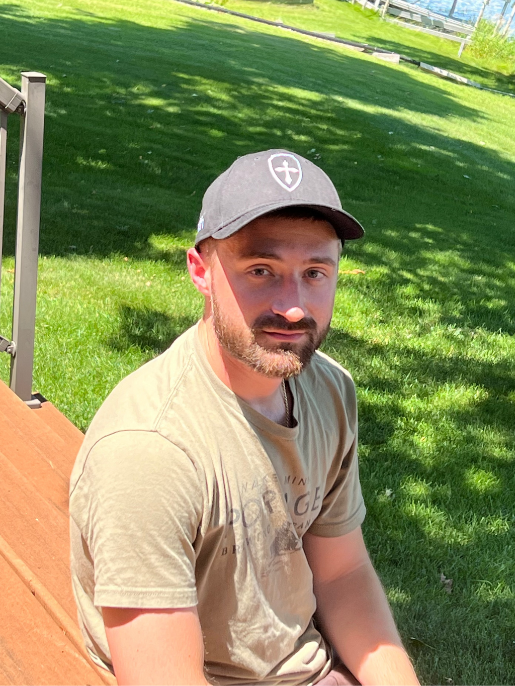 A man wearing a black baseball cap with a white cross logo and a beige t-shirt, sitting outdoors on a wooden bench in a grassy park area.