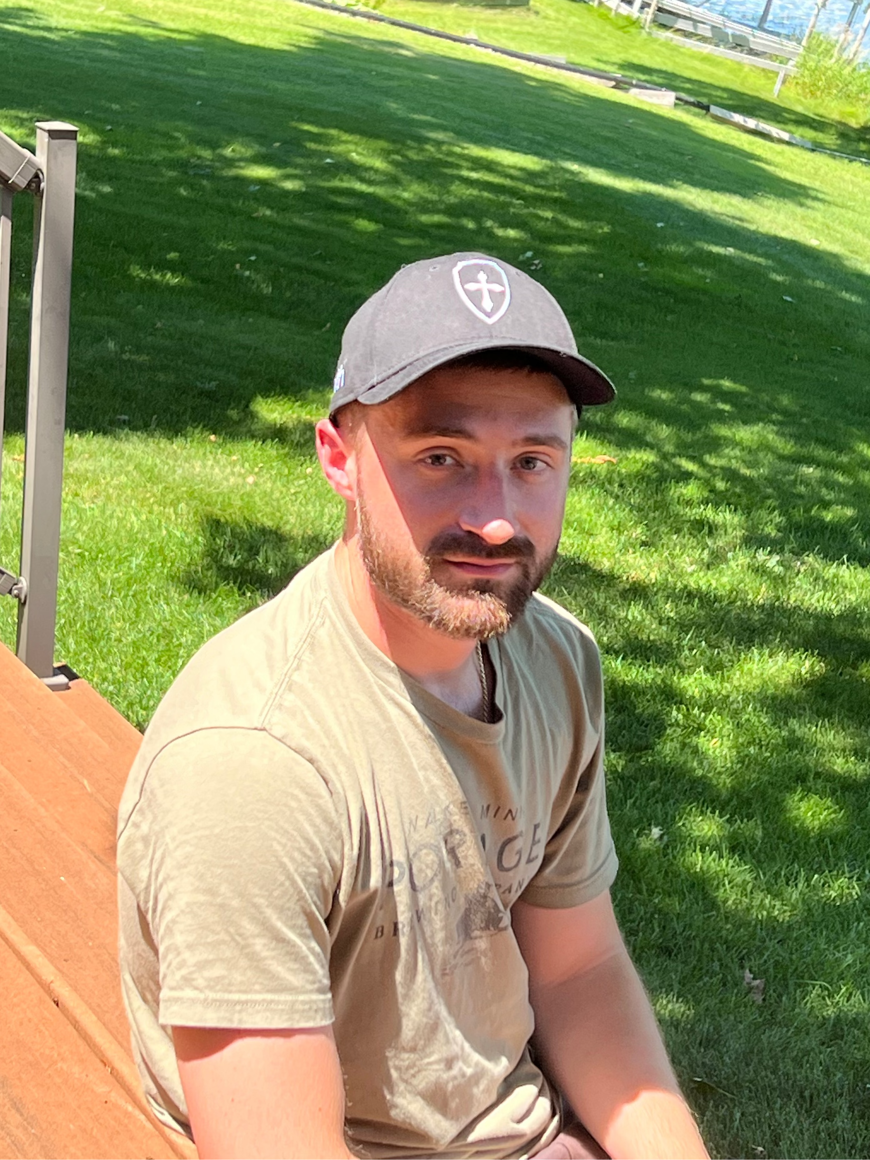 A man wearing a black baseball cap with a white cross logo and a beige t-shirt, sitting outdoors on a wooden bench in a grassy park area.